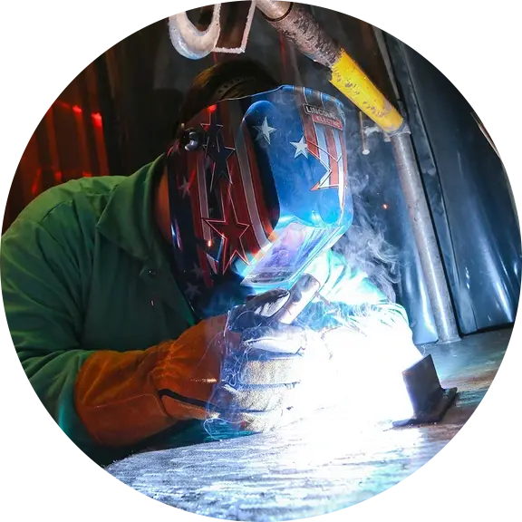 Student wearing a patriotic welding helmet and protective gear while welding metal, surrounded by sparks and smoke.