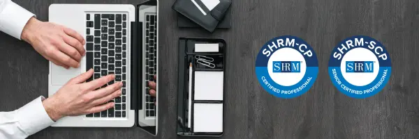 Top-down view of a person typing on a laptop next to a notepad and pen tray on a dark wood desk, with SHRM-CP and SHRM-SCP certification badges displayed to the right.
