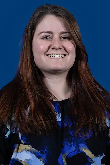 Portrait of a smiling woman with long brown hair, wearing a colorful blouse with blue, black, and yellow patterns, posed in front of a solid blue background.