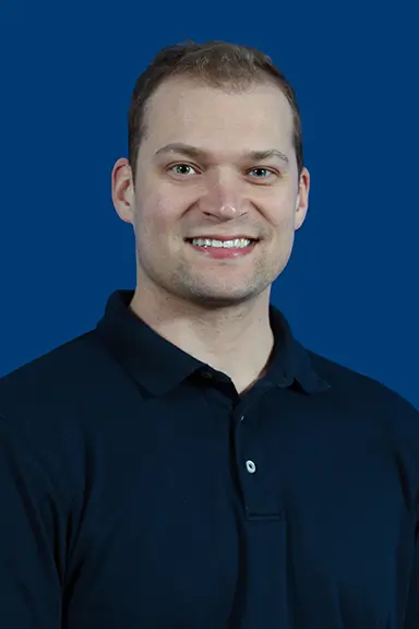 Portrait of a smiling man with short brown hair, wearing a dark navy polo shirt, posed in front of a solid blue background.