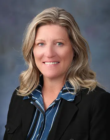Professional headshot of a woman with blonde, shoulder-length wavy hair, wearing a blue striped blouse and a black blazer, smiling in front of a soft, gray-blue background.