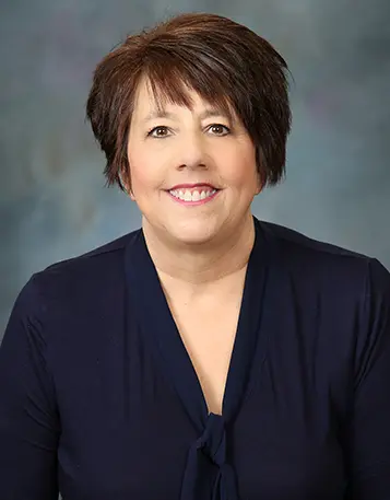 Professional headshot of a woman with short dark brown hair and side-swept bangs, smiling, wearing a navy top, and posed against a neutral blue-gray background.