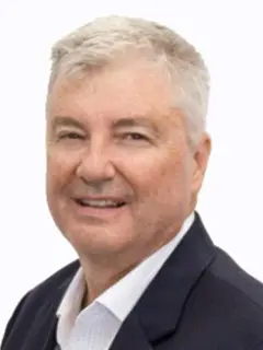 Professional headshot of a smiling man with short gray hair, wearing a dark suit jacket over a white collared shirt, posed against a light background.