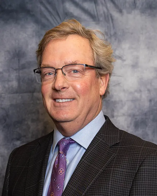 Portrait of a man with light hair and glasses wearing a dark plaid suit, light blue dress shirt, and patterned purple tie, standing in front of a mottled gray backdrop.