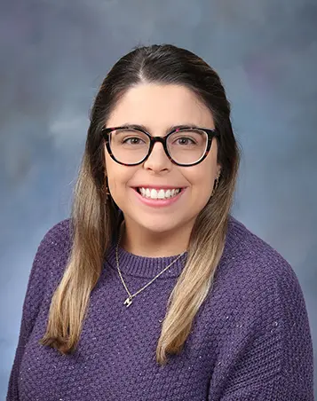 Professional headshot of a woman with long, straight dark blonde hair, wearing glasses and a purple sweater, smiling in front of a soft blue studio background.