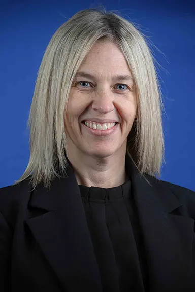 Professional headshot of a woman with straight, shoulder-length blonde hair, wearing a black blazer and black blouse, smiling in front of a solid blue background.