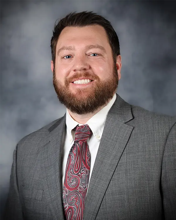 Portrait of a man with short dark hair and a full beard, wearing a gray suit jacket, white dress shirt, and a red paisley tie, smiling in front of a neutral gray background