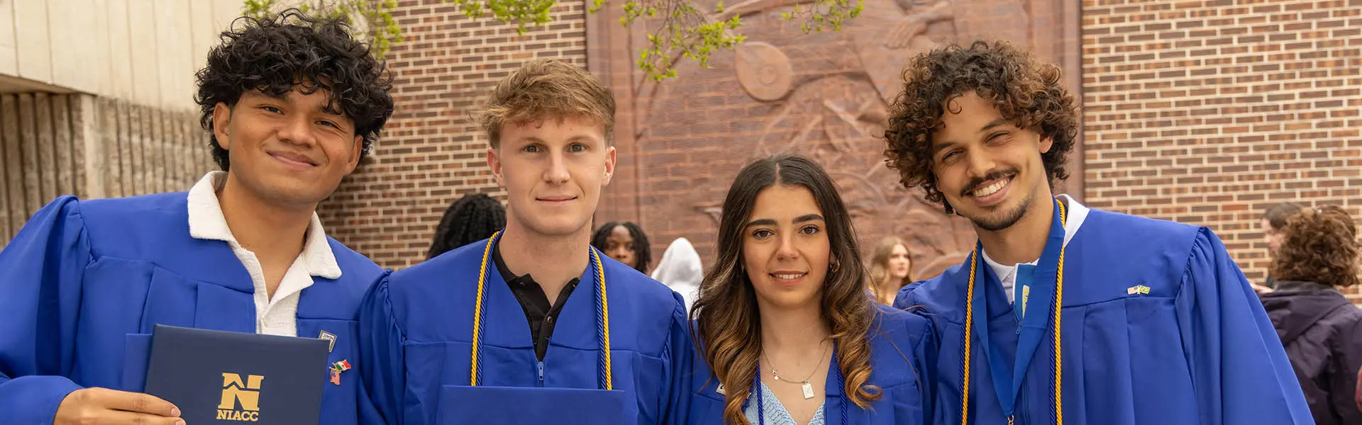 Four NIACC graduates in blue caps and gowns stand together smiling and holding diplomas during a graduation ceremony.