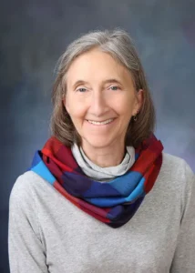 Woman with gray hair wearing a scarf smiles at the camera in a studio-style professional headshot.