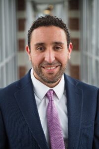 Man in a suit and tie smiling at the camera in a professional headshot taken in a hallway.