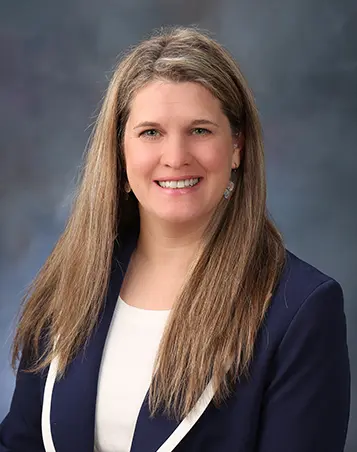 Professional headshot of a woman with long light brown hair wearing a navy blazer and white top, smiling against a gray studio background.