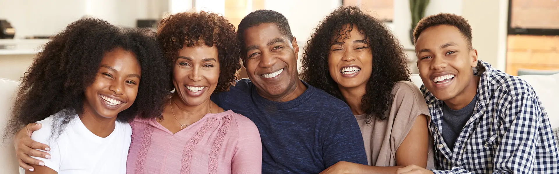 Smiling family of five&mdash;two adults and three young adults&mdash;sitting close together indoors, conveying warmth and connection.