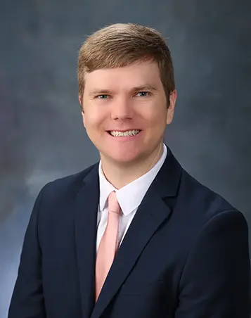 Professional headshot of a man in a navy suit and light pink tie, smiling against a gray studio background.