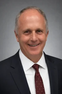 Professional headshot of a man wearing a dark suit, white shirt, and burgundy tie, smiling against a gray background.