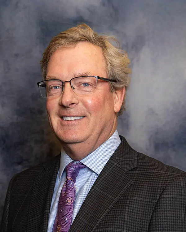 Portrait of a man with light hair and glasses wearing a dark plaid suit, light blue dress shirt, and patterned purple tie, standing in front of a mottled gray backdrop.