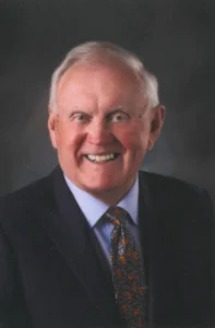 Professional portrait of an older man wearing a dark suit and patterned tie, smiling against a dark studio background.