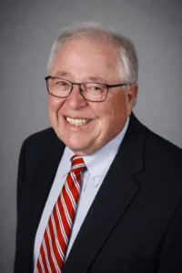 Professional headshot of an older man wearing glasses, a dark suit, and a red striped tie, smiling against a gray background.