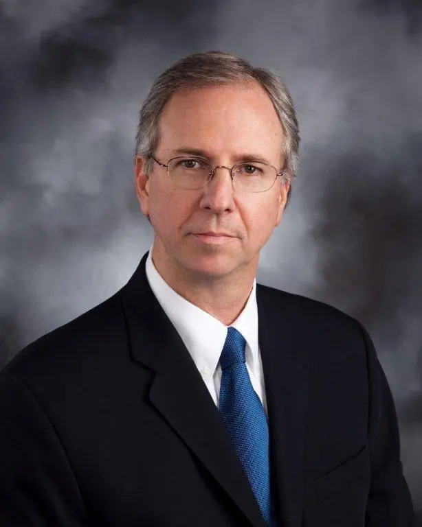 Man with short gray hair and glasses wearing a dark suit and blue tie poses for a professional portrait against a gray background.