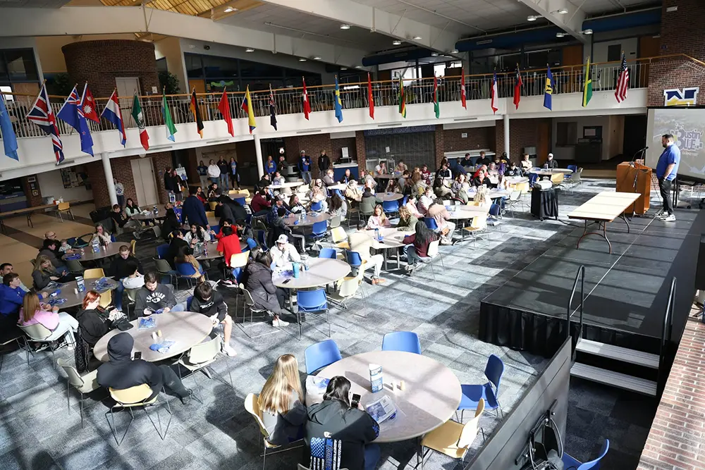 Students gather around tables in the NIACC Activity Center while a speaker presents from a stage beneath a display of international flags.