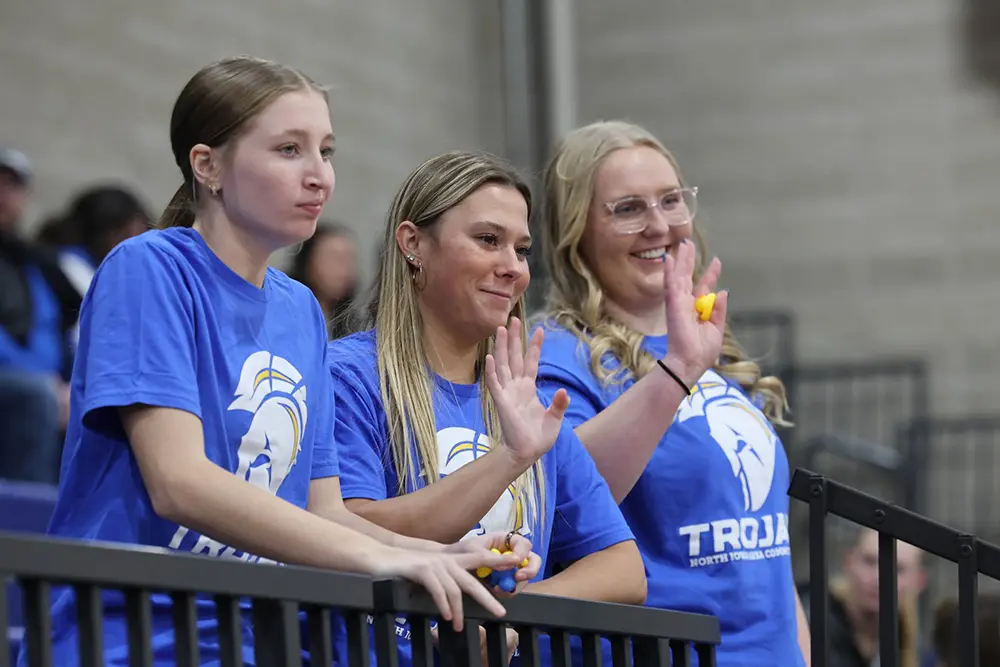 Three NIACC students in Trojan T-shirts stand in the gymnasium bleachers, two waving toward the court.