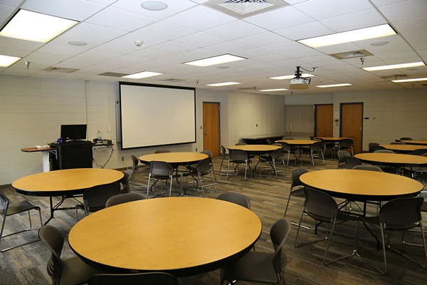Photo of meeting room AC101 showing tables, chairs, projector, projector screen, and command station